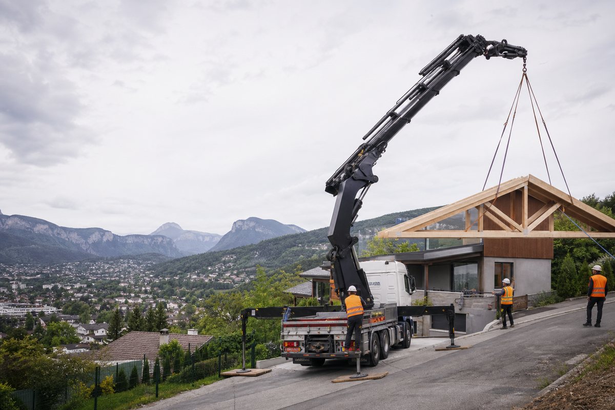 Mise en situation d’un camion-grue européen avec une équipe de levage pour charpente de chalet sur versant, dans un environnement inspiré de Chambéry